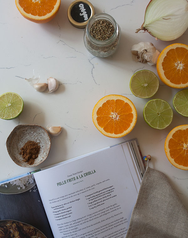 Cut of oranges, limes, and spices on a table with an open cookbook near by.