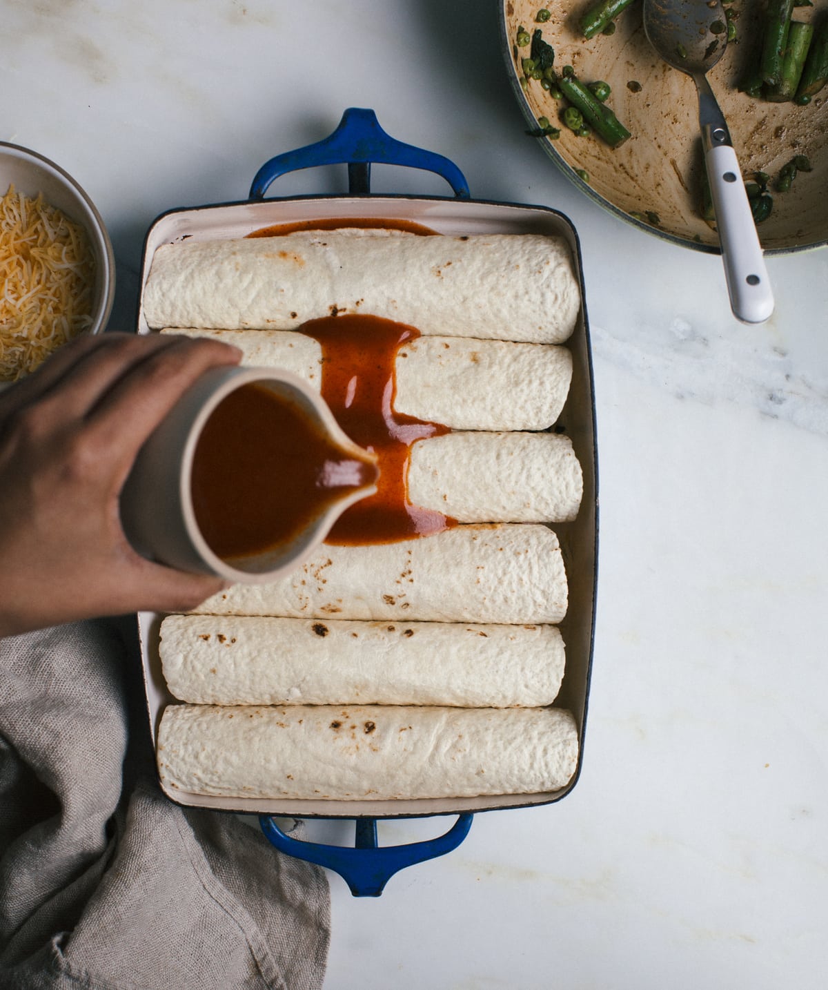 Hand pouring enchilada sauce over enchiladas in a pan. 