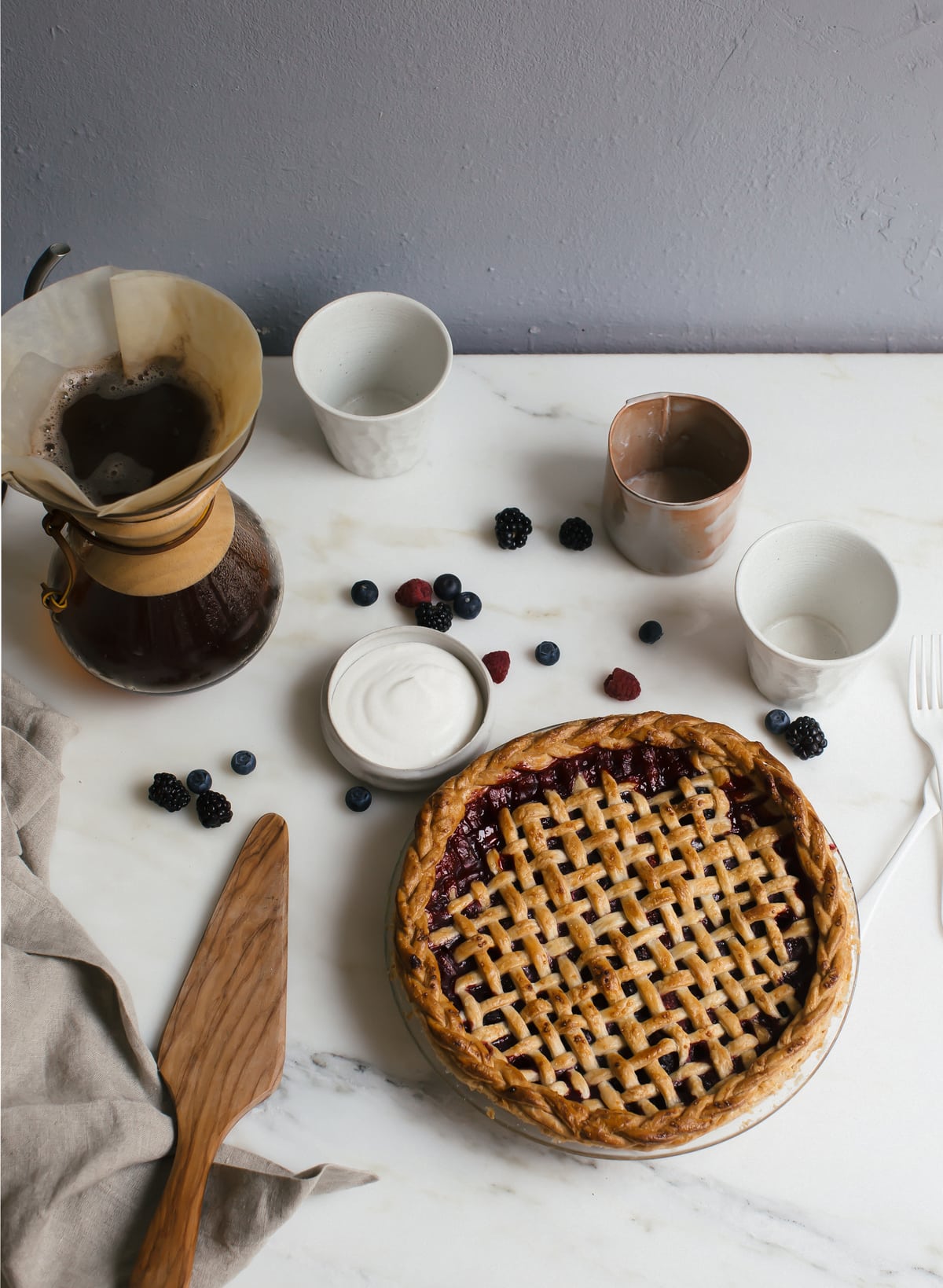 Baked mixed berry pie on a counter with coffee and fresh fruit near by.
