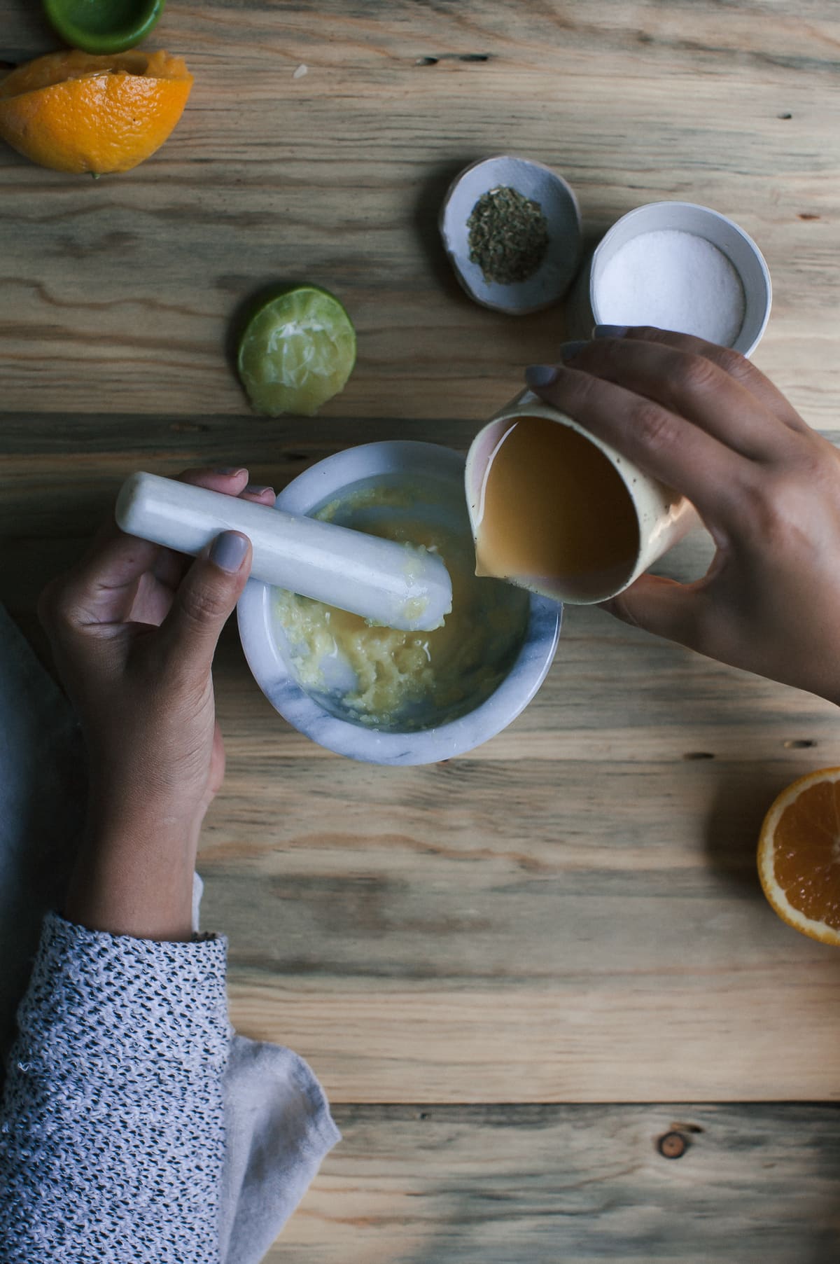 Mashed garlic in a mortar with hands holding the pestle and pouring in liquid into the mortar.