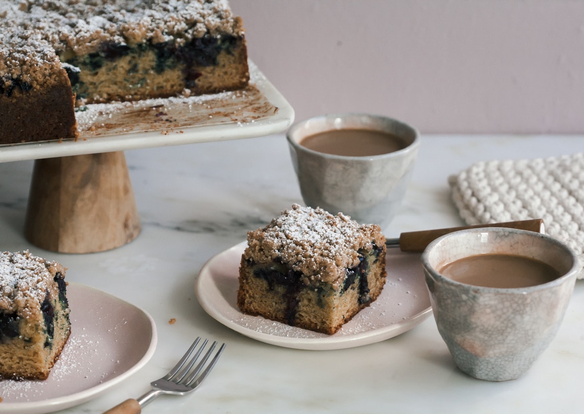 Close up image of blueberry coffee cake on a plate with a cup of coffee near by. 