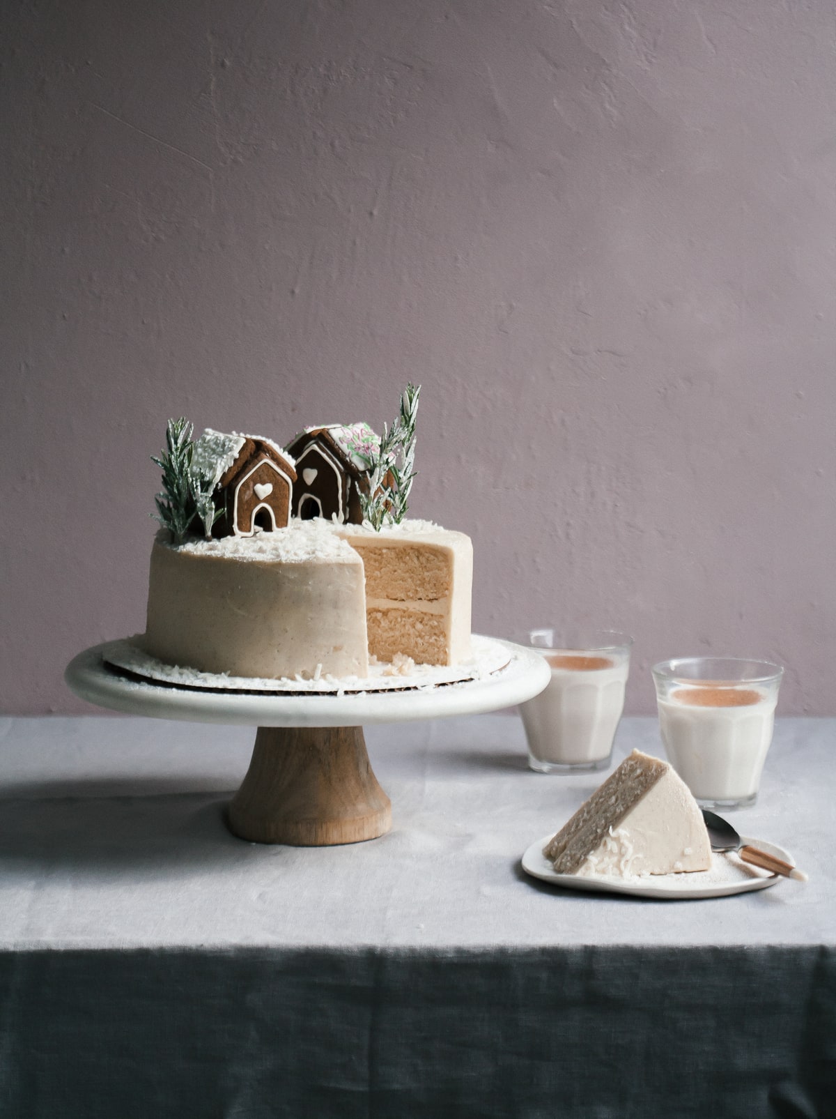 Horchata cake topped with gingerbread houses on a cake stand with a slice of cake on a plate nearby.