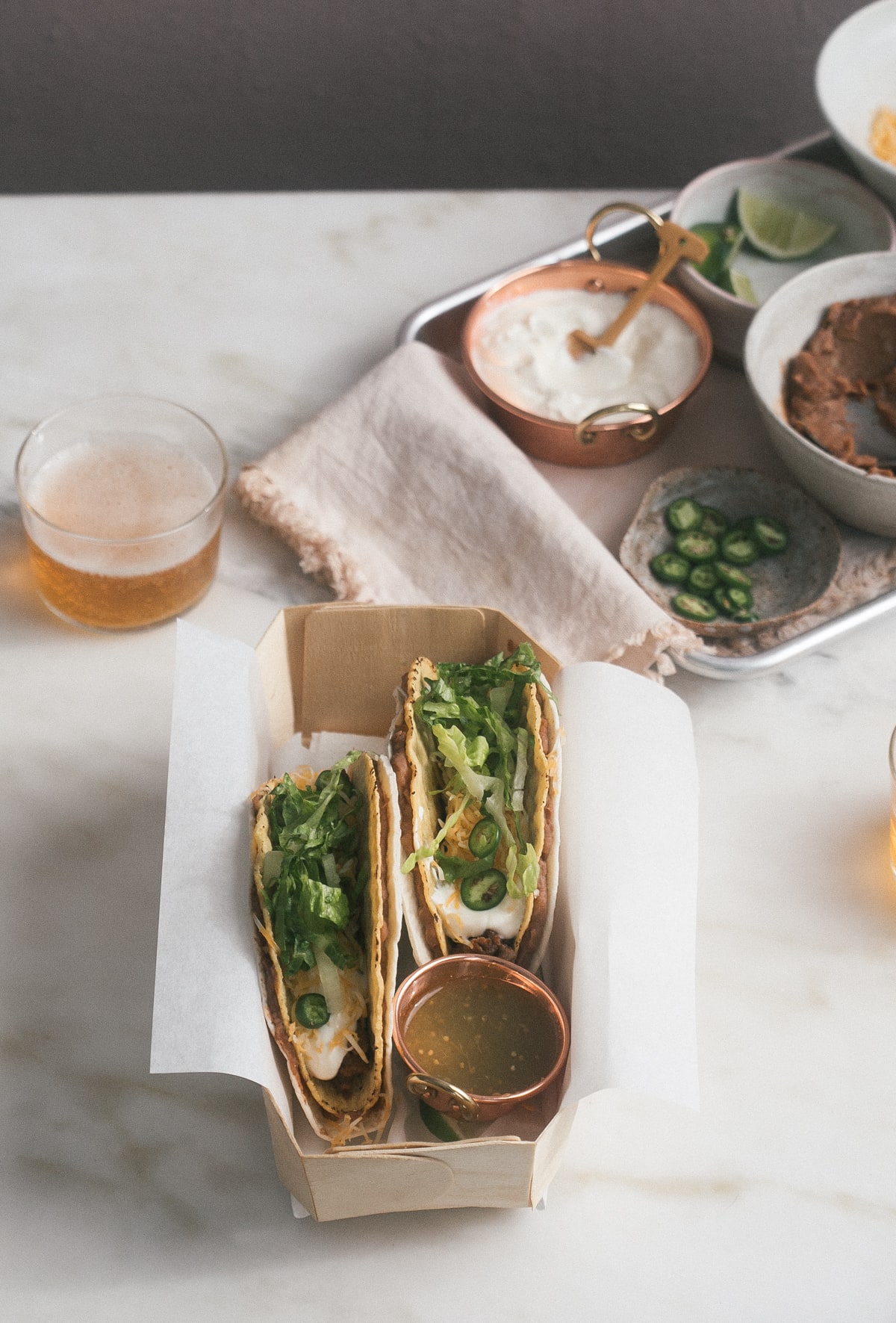 3/4 image of tacos in a box with a glass of beer and taco toppings near by.