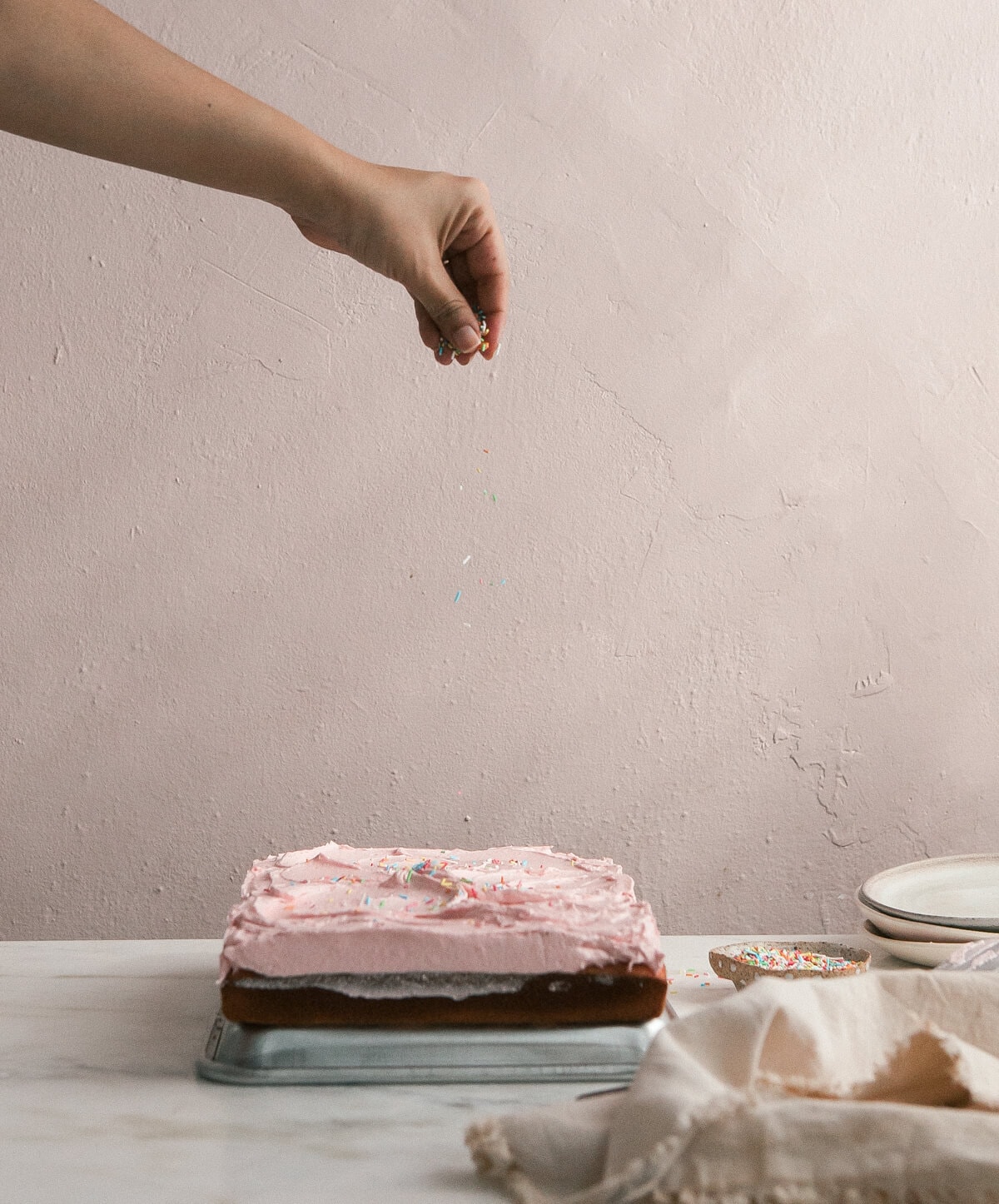 A hand sprinkling sprinkles over a frosted cake. 