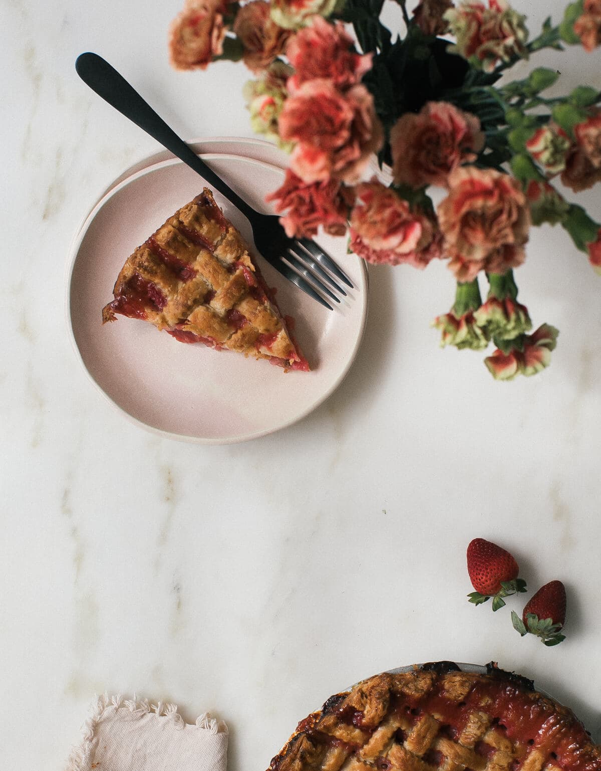 Overhead image of a slice of rhubarb strawberry pie on a plate with a fork and flowers near by.
