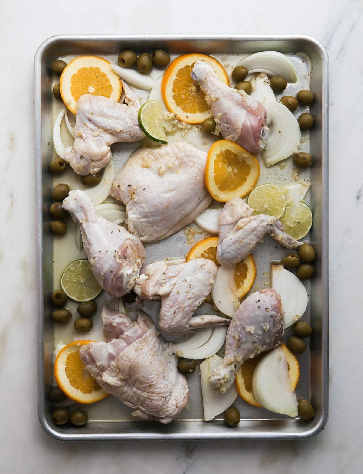 Raw chicken, orange and lime slices, onions and olives arranged on a sheet pan.