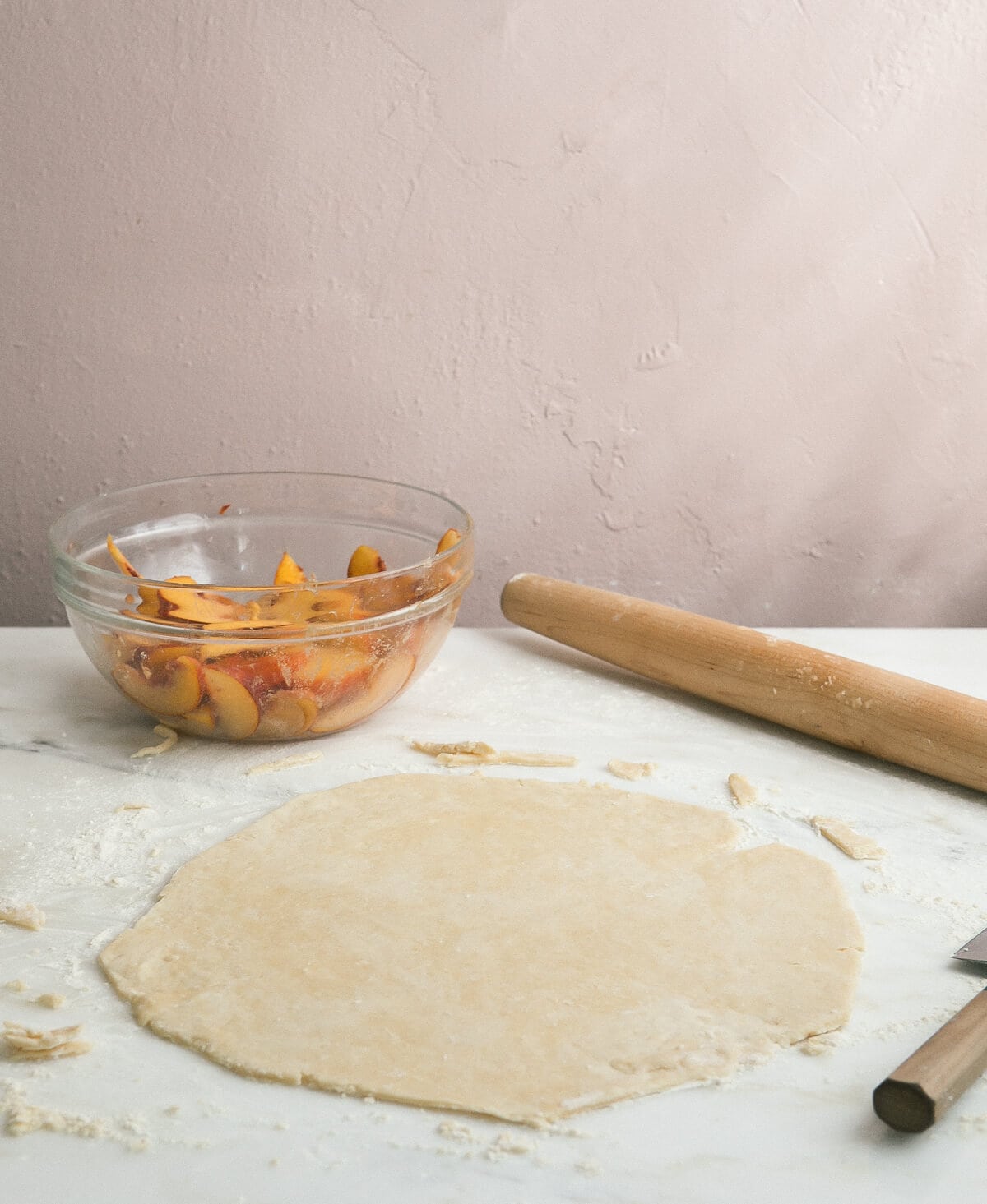 Pie crust rolled out with a rolling pin and a bowl of sliced peaches near by.