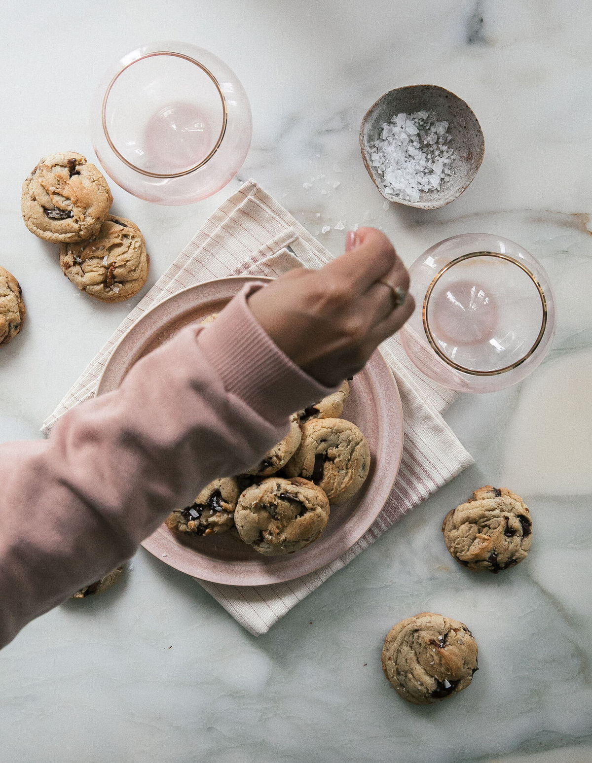 Hand sprinkling flaky salt over a plate of bourbon pecan chocolate chip cookies.