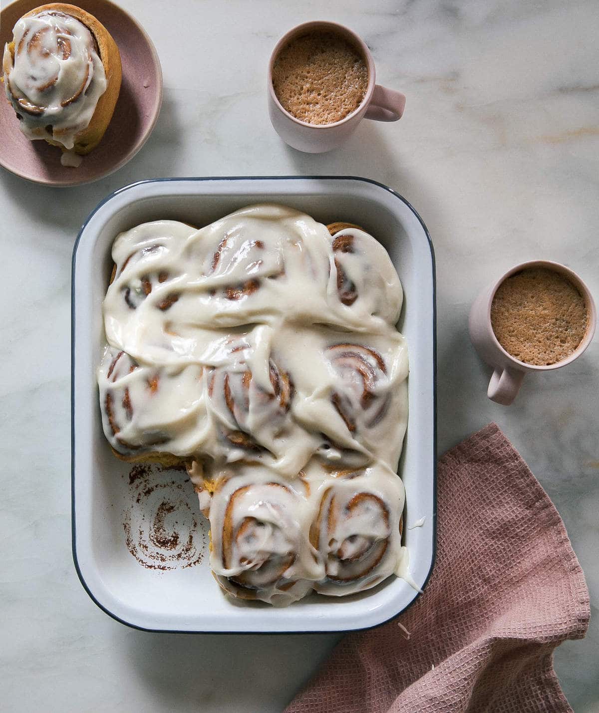 Cinnamon rols with maple cream cheese frosting on top. Surrounded by coffee cups and plates.