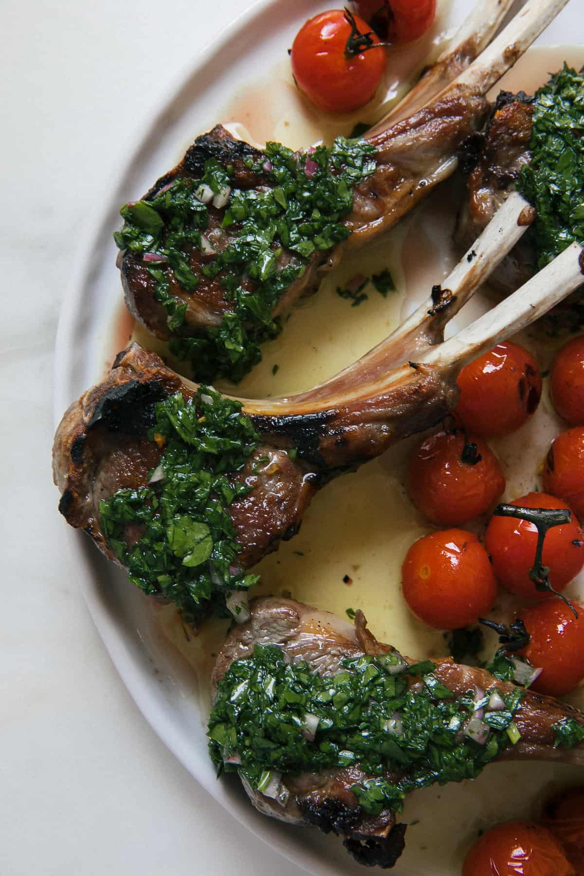 Lamb chops on plate with chimichurri and roasted tomatoes. 