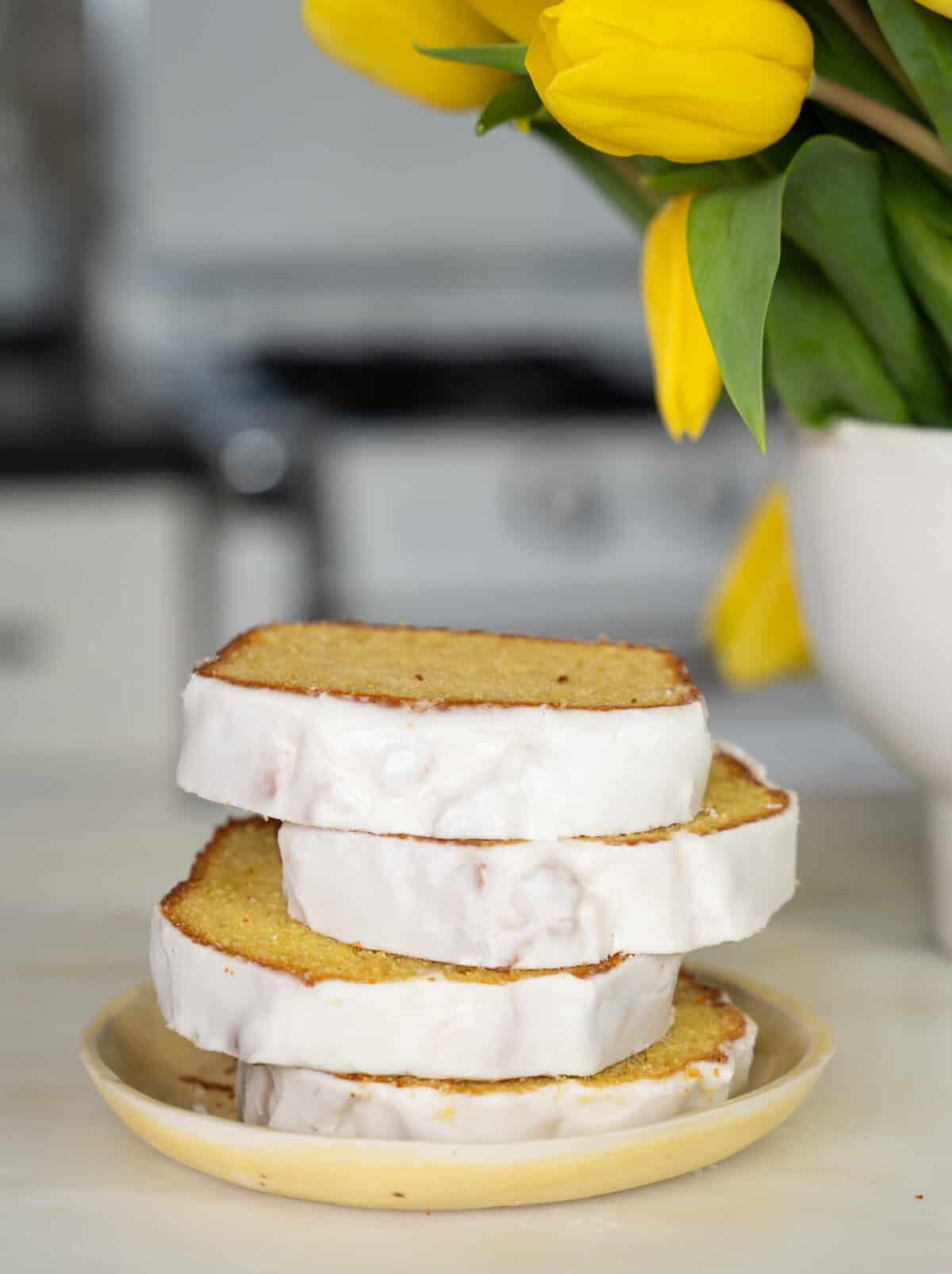 Slices of lemon loaf on top of each other on a yellow plate with tulips in the background. 