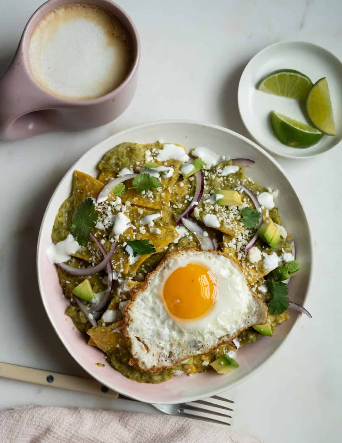 Chilaquiles Verdes in a bowl with a fried egg on top. Coffee and lime slices are on the side. 