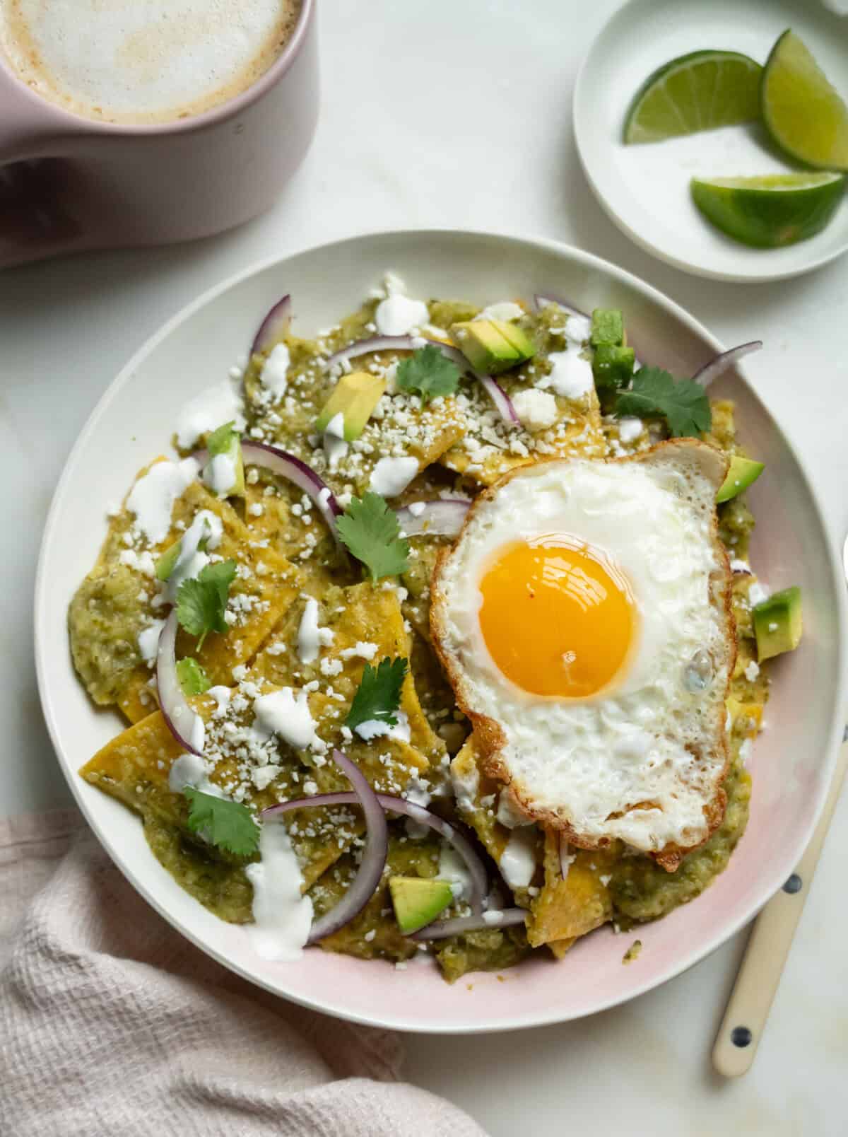 Chilaquiles Verdes in a bowl with a fried egg on top. Limes on the side with a cup of coffee. 