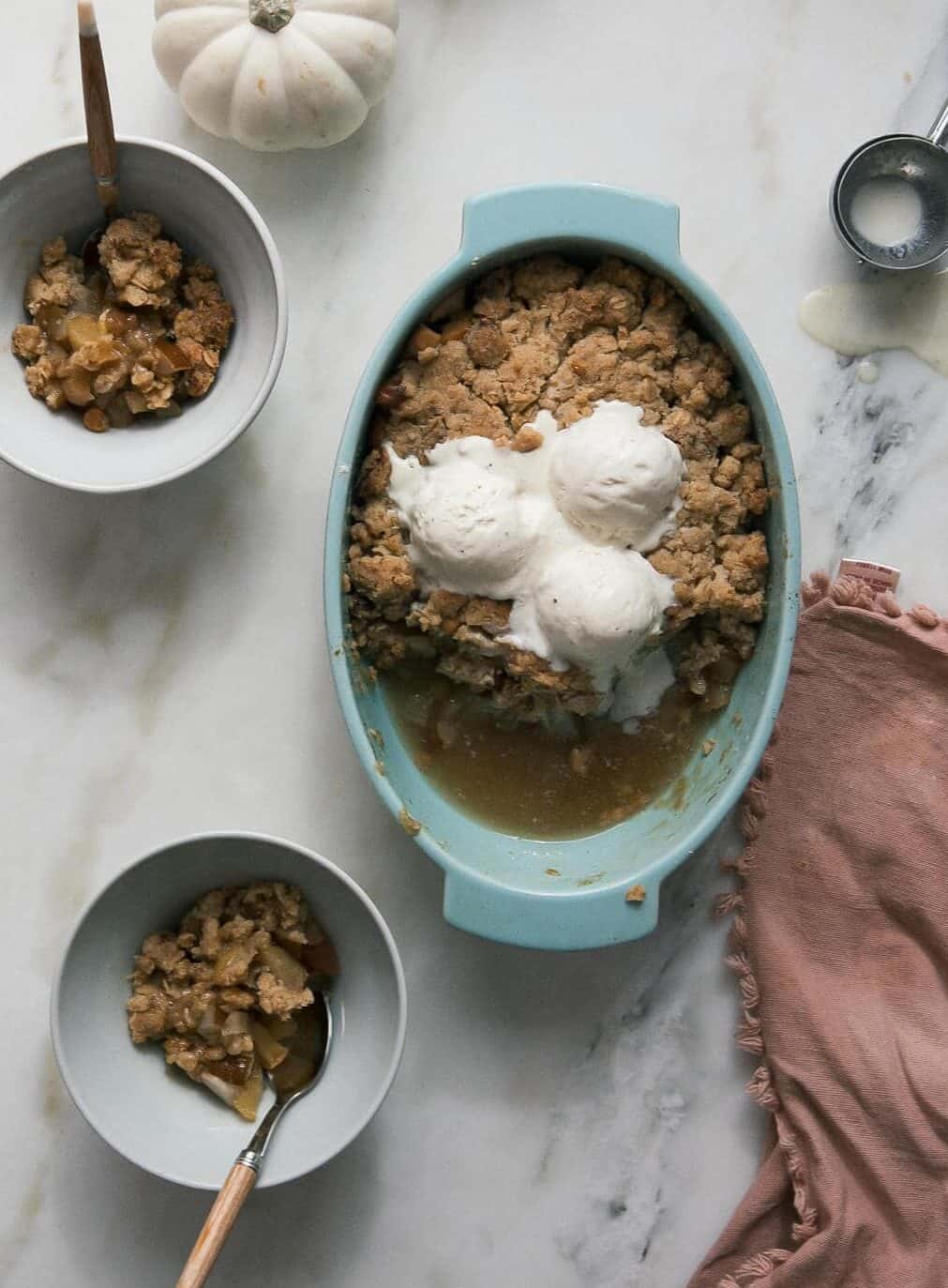 A casserole of brown butter pear apple crumble with three scoops of vanilla ice cream and two bowls of brown butter pear apple crumble.