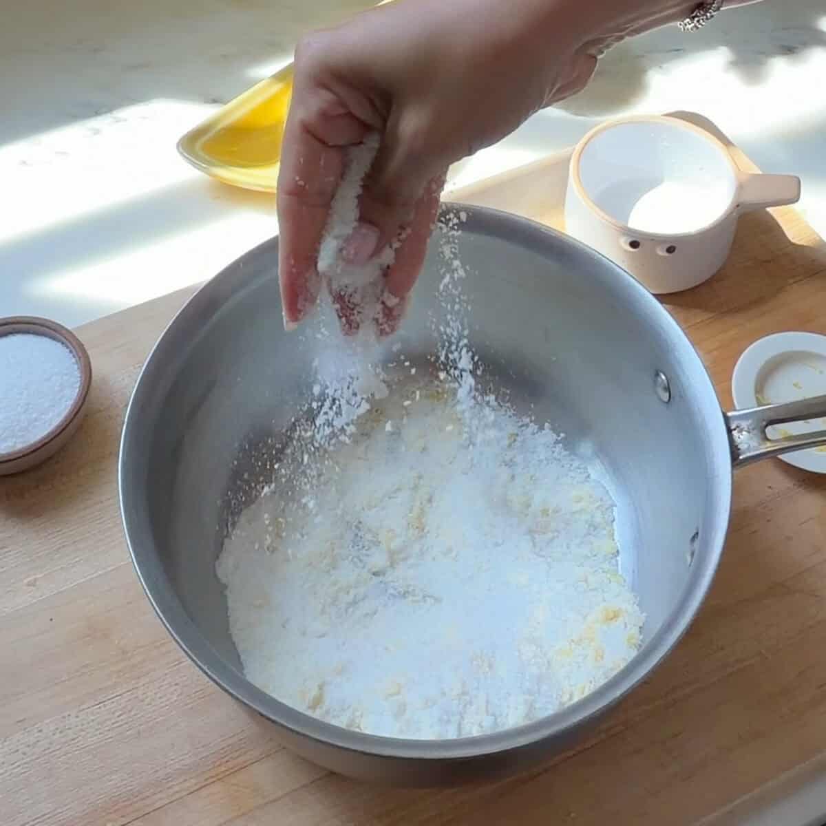 Fingers rubbing lemon zest into sugar in pot.