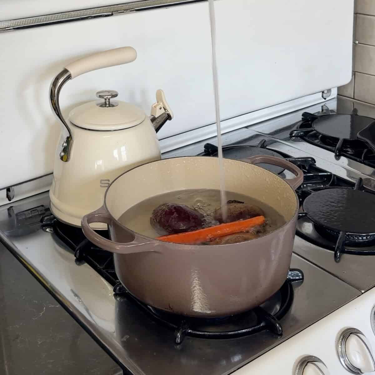 Adding water to a large pot with carrot, beets, and potatoes in it.