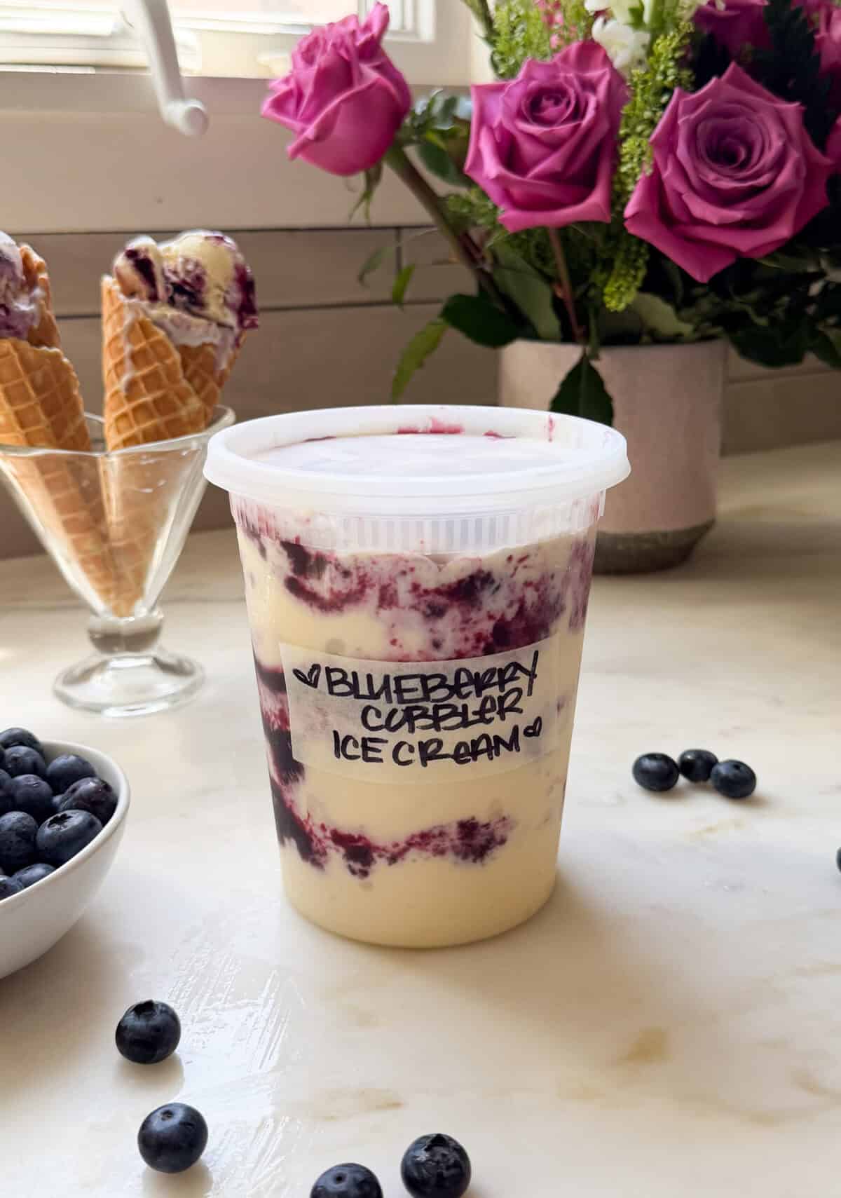 Quart deli container filled with Blueberry Cobbler Ice Cream and ice cream melty ice cream cones in the background.