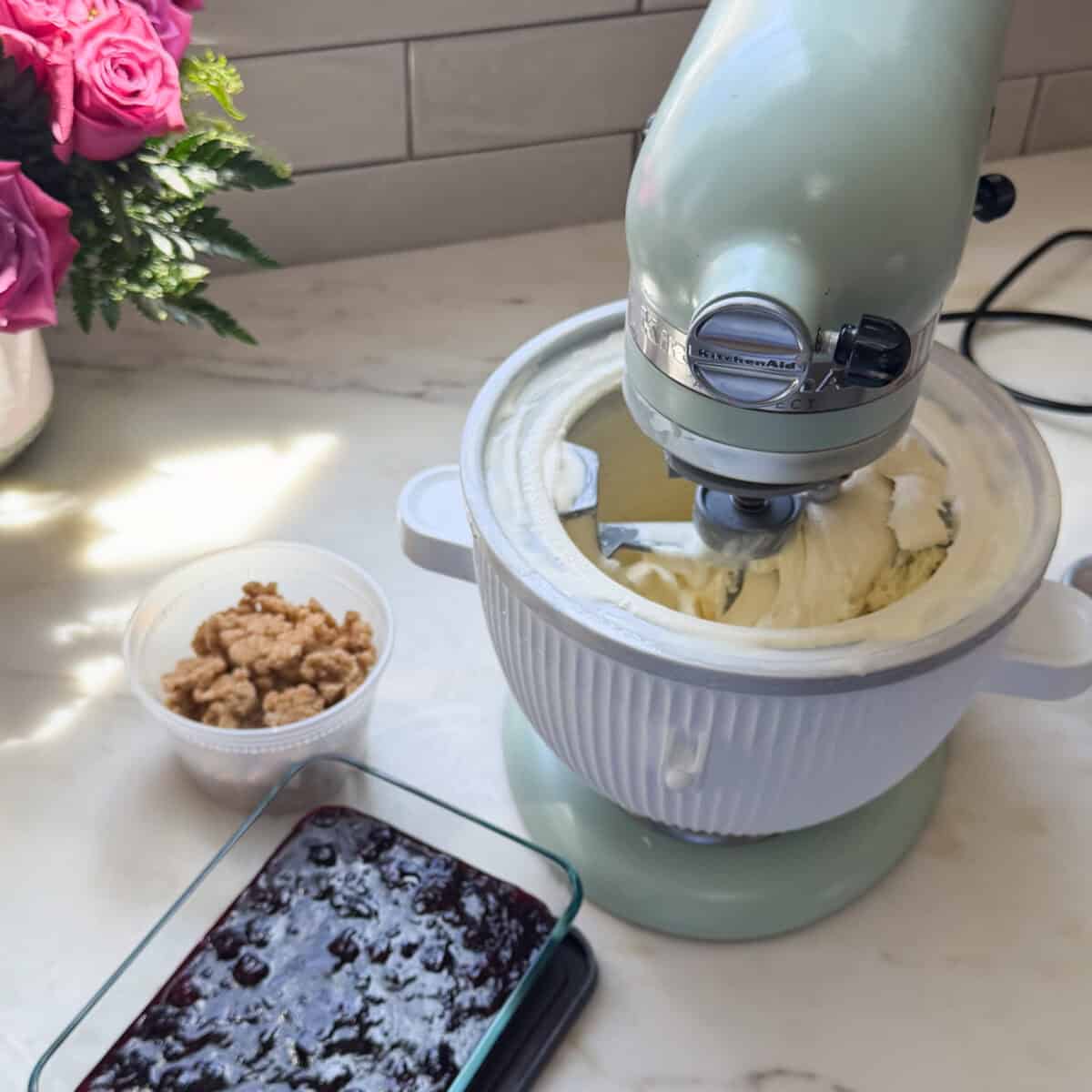 Stand mixer mixing vanilla ice cream, a container of blueberry compote, and a deli container of crumble.