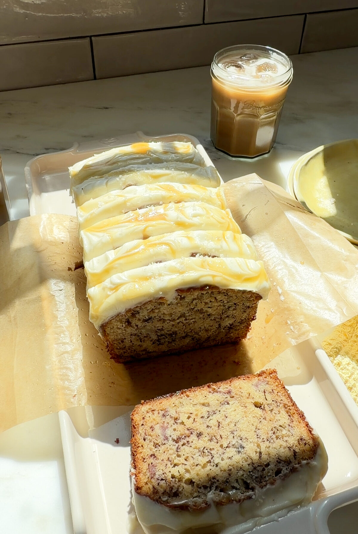 Sliced and frosted salted maple banana bread loaf on a platter with an iced coffee.